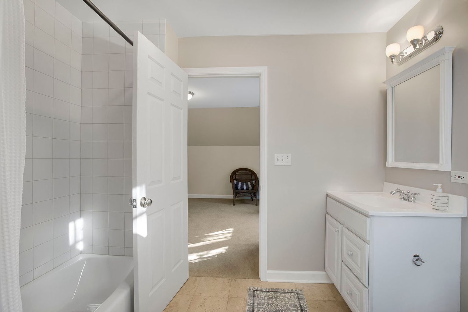 This is a bright and clean bathroom featuring a white vanity with a sink, a mirror with overhead lighting, and a tiled shower/tub combination. The bathroom has a neutral color palette with beige walls and tile flooring, and an open doorway leads to another room with a wicker chair, suggesting a guest bedroom or attic space. The overall impression is neat and well-maintained, suitable for a guest or secondary bathroom.