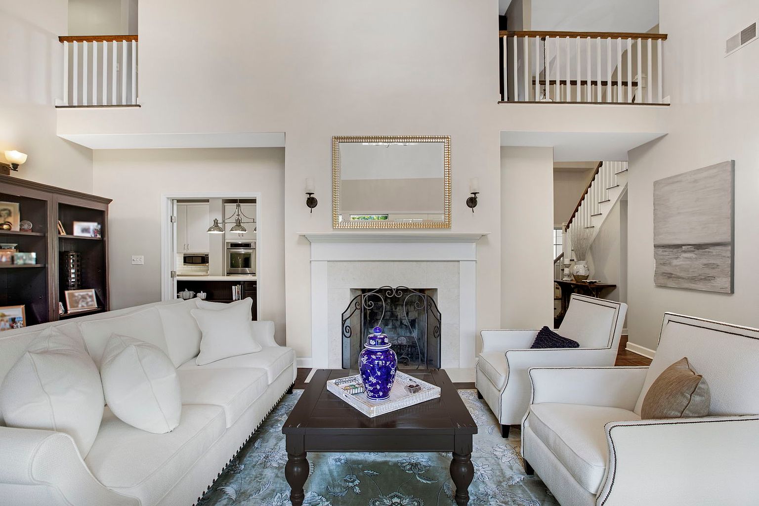 This is an interior shot of a living room featuring a white sofa and two white armchairs arranged around a dark wood coffee table. A fireplace with a decorative screen and a gold-framed mirror above it serves as a focal point. The room has high ceilings and a neutral color palette, creating a bright and airy atmosphere.