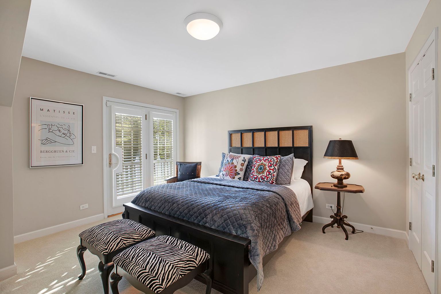 The image showcases a well-lit guest bedroom featuring a dark wood bed frame with a woven headboard, complemented by a blue quilted comforter and decorative pillows. Two zebra-striped benches sit at the foot of the bed, and a framed Matisse print hangs on the wall. A small side table with a lamp adds a cozy touch, while a door leads to a balcony, enhancing the room's appeal.