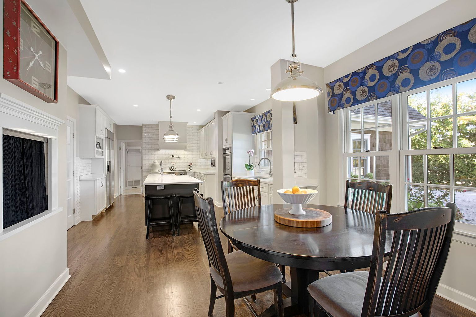 This is a bright and airy kitchen and dining area featuring hardwood floors and white cabinetry. A round wooden dining table with chairs sits in the foreground, illuminated by a pendant light, while a kitchen island with bar stools is visible in the background. The space is well-lit with natural light streaming through a large window, creating a warm and inviting atmosphere.