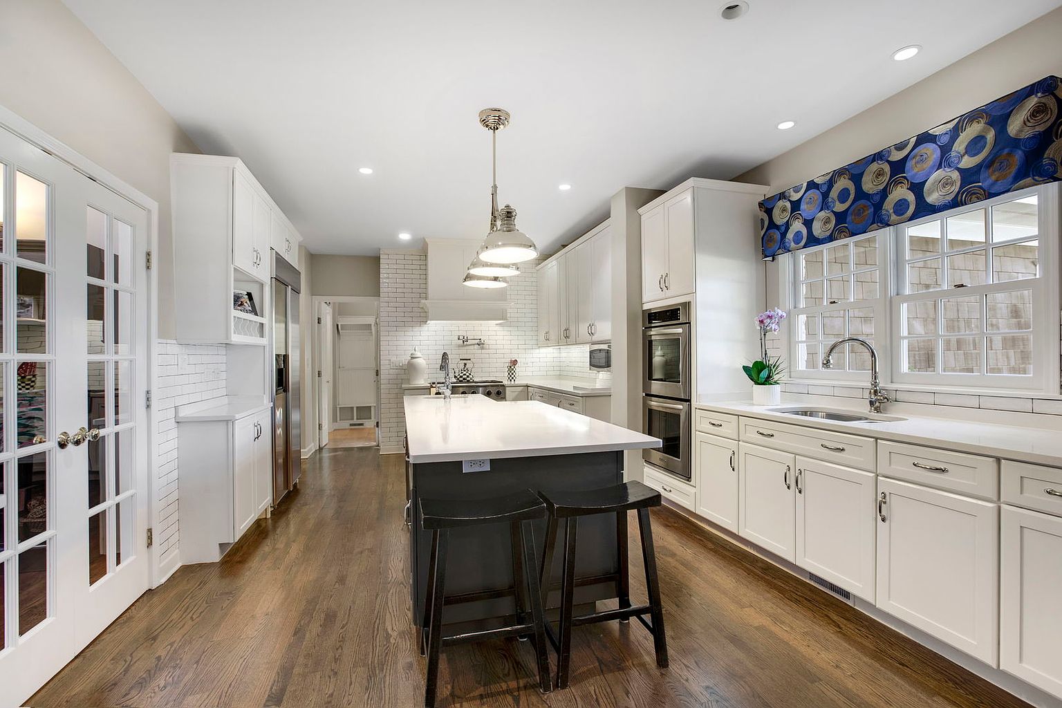 This is a bright and spacious kitchen featuring white cabinetry, stainless steel appliances, and a dark-colored island with a white countertop. The kitchen has hardwood floors and a window with a patterned valance. The perspective is from the entrance of the kitchen, showcasing the island and the appliances.