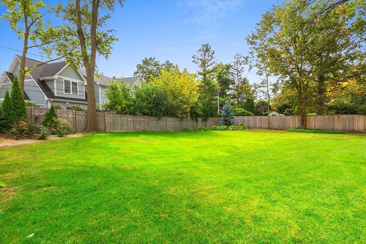 The image showcases a spacious, well-maintained backyard with a lush green lawn. A wooden fence encloses the yard, providing privacy, while mature trees and landscaping add to the aesthetic appeal. A portion of a house is visible on the left, suggesting a residential setting.