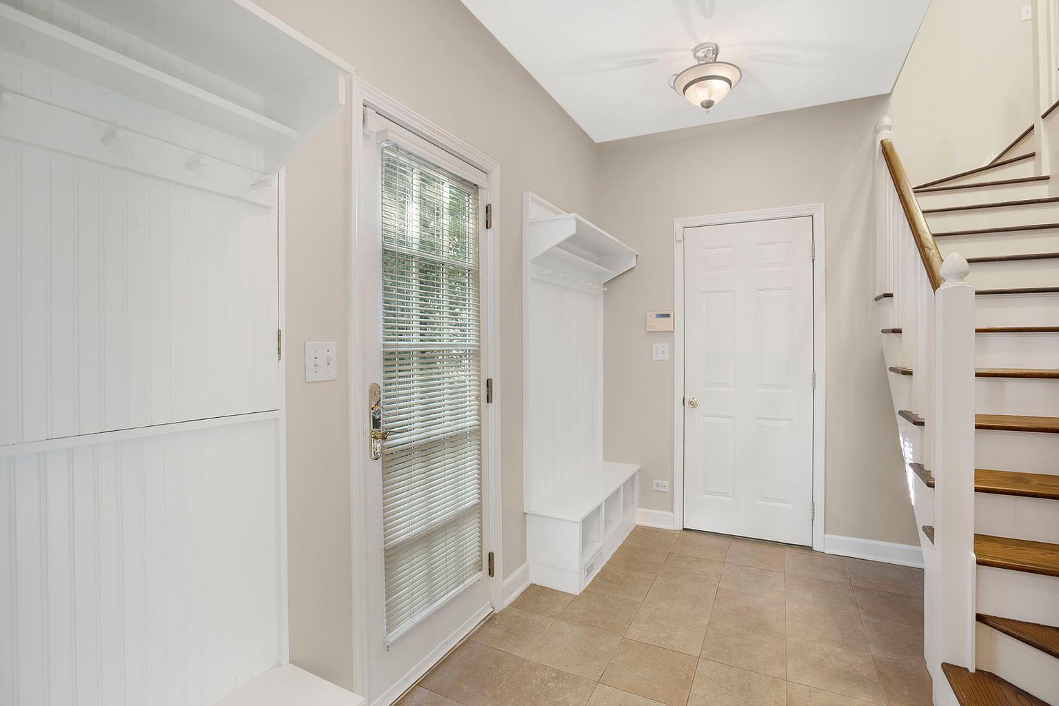 This interior shot showcases a well-lit hallway with neutral-toned walls and tiled flooring. A white staircase with wooden treads is visible on the right, while a built-in storage unit and a door with blinds are on the left. A coat rack and another door are visible straight ahead, creating a functional and inviting entryway.