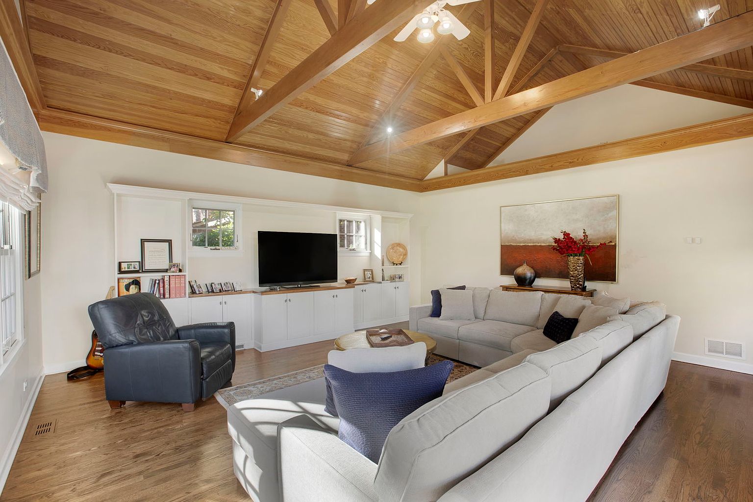 This is a well-lit living room featuring a high, vaulted ceiling with exposed wooden beams and a wood plank ceiling. A large sectional sofa anchors the space, complemented by a leather armchair and a built-in entertainment center with a flat-screen TV. The room is decorated with neutral tones and a large piece of artwork, creating a warm and inviting atmosphere.