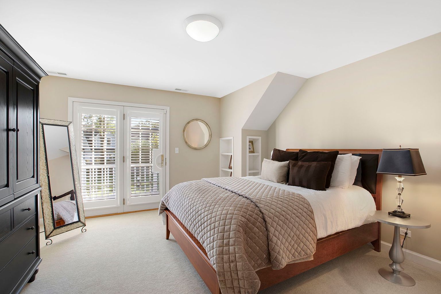This is a well-lit primary bedroom featuring a bed with a quilted coverlet and several pillows, a dark wood armoire, and a full-length mirror. French doors with white shutters lead to the outside, and a round mirror hangs on the wall. The room has a cozy and inviting atmosphere, perfect for relaxation.