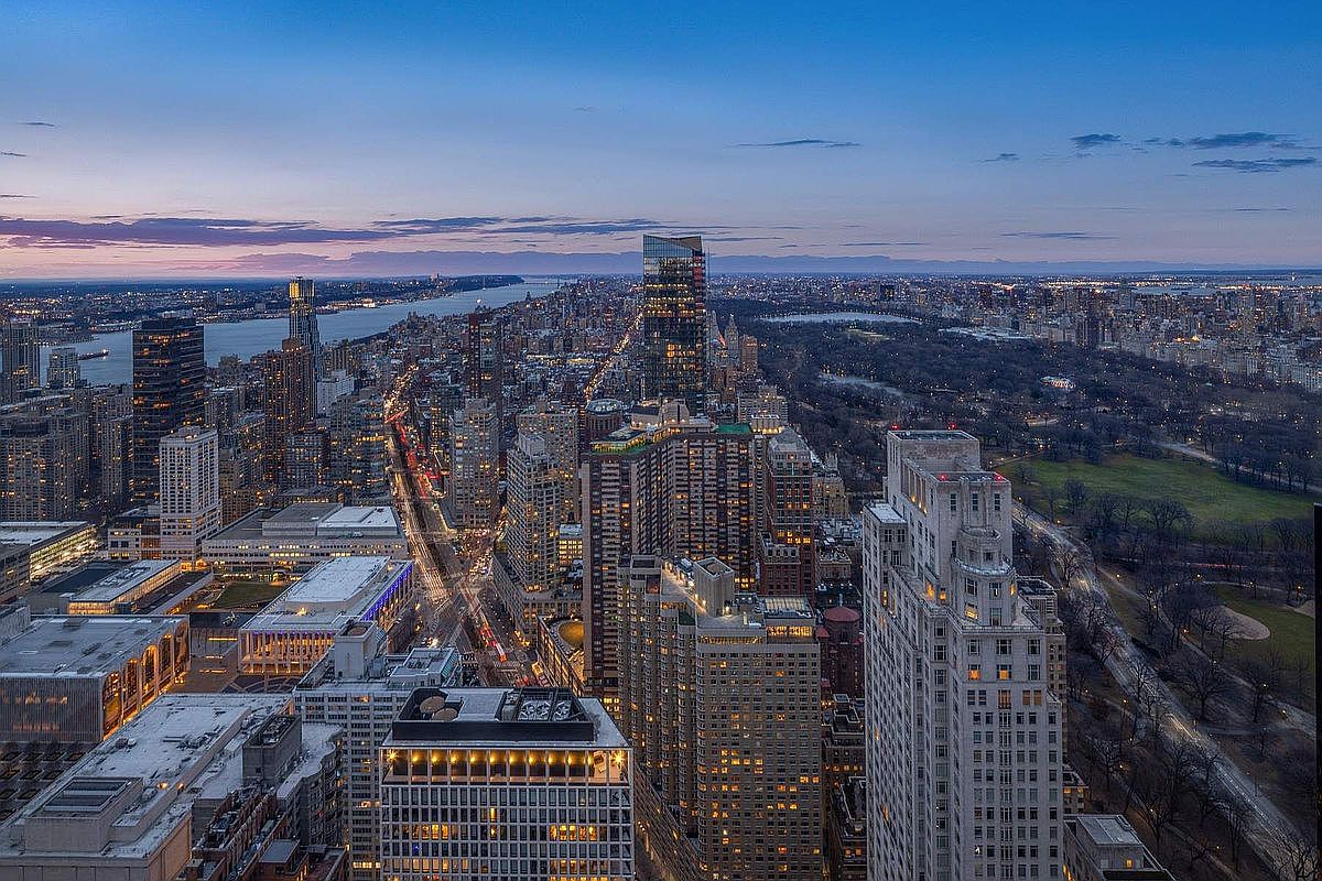 This high-altitude aerial view captures a breathtaking twilight panorama of the New York City skyline, prominently featuring the lush expanse of Central Park adjacent to dense urban architecture. The scene highlights the contrast between the illuminated skyscrapers and the darkening park, with the Hudson River visible in the distance under a soft, gradient sunset sky. The perspective offers a cinematic sense of scale, emphasizing the prestigious and vibrant location of the property within the heart of Manhattan.