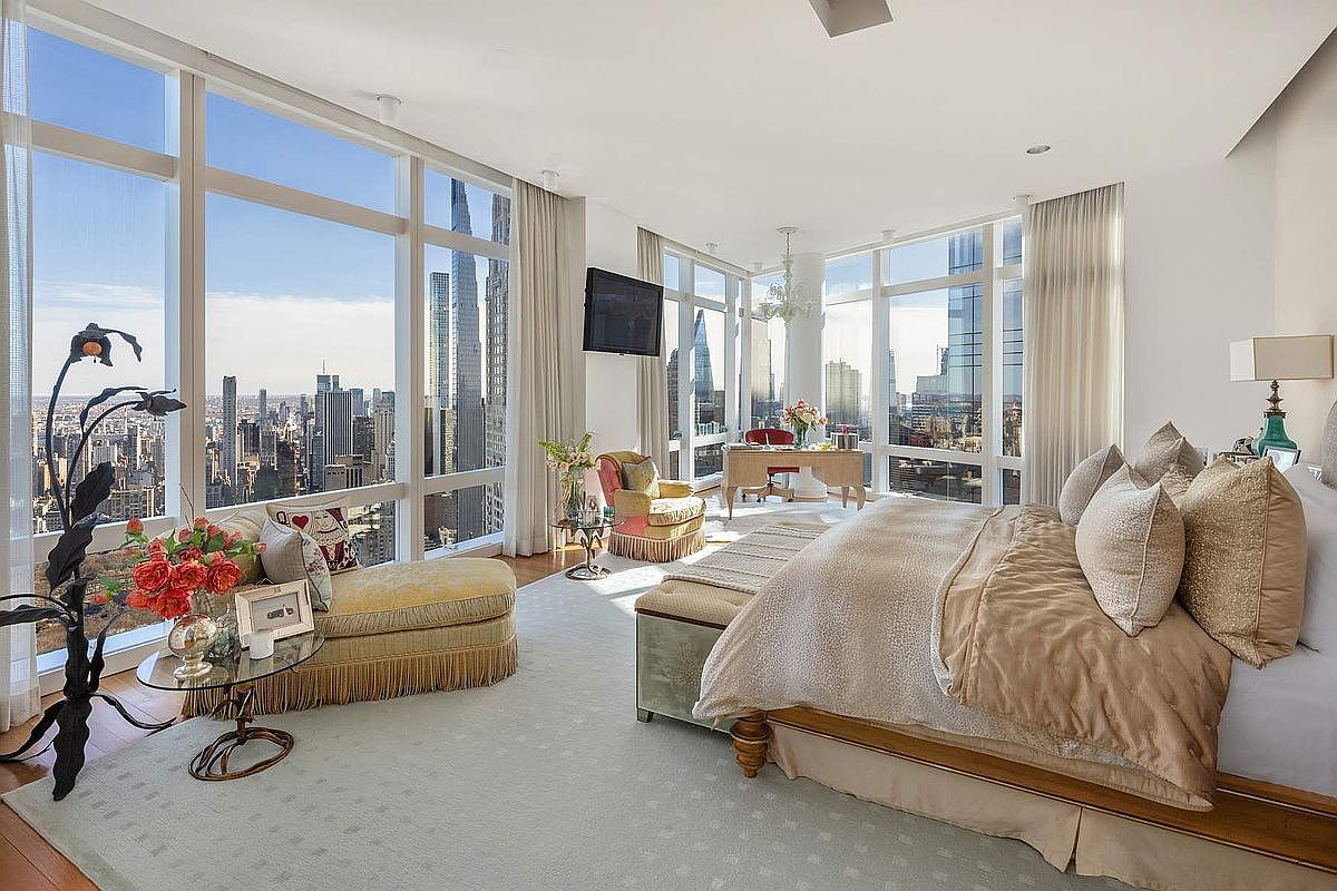 This luxurious primary bedroom features floor-to-ceiling windows that offer a breathtaking, panoramic view of the New York City skyline. The room is elegantly furnished with a plush king-sized bed, a sophisticated chaise lounge, and a dedicated workspace, all set against a neutral, high-end color palette. The combination of natural light, opulent textures, and an iconic urban backdrop creates an atmosphere of refined, elevated city living.