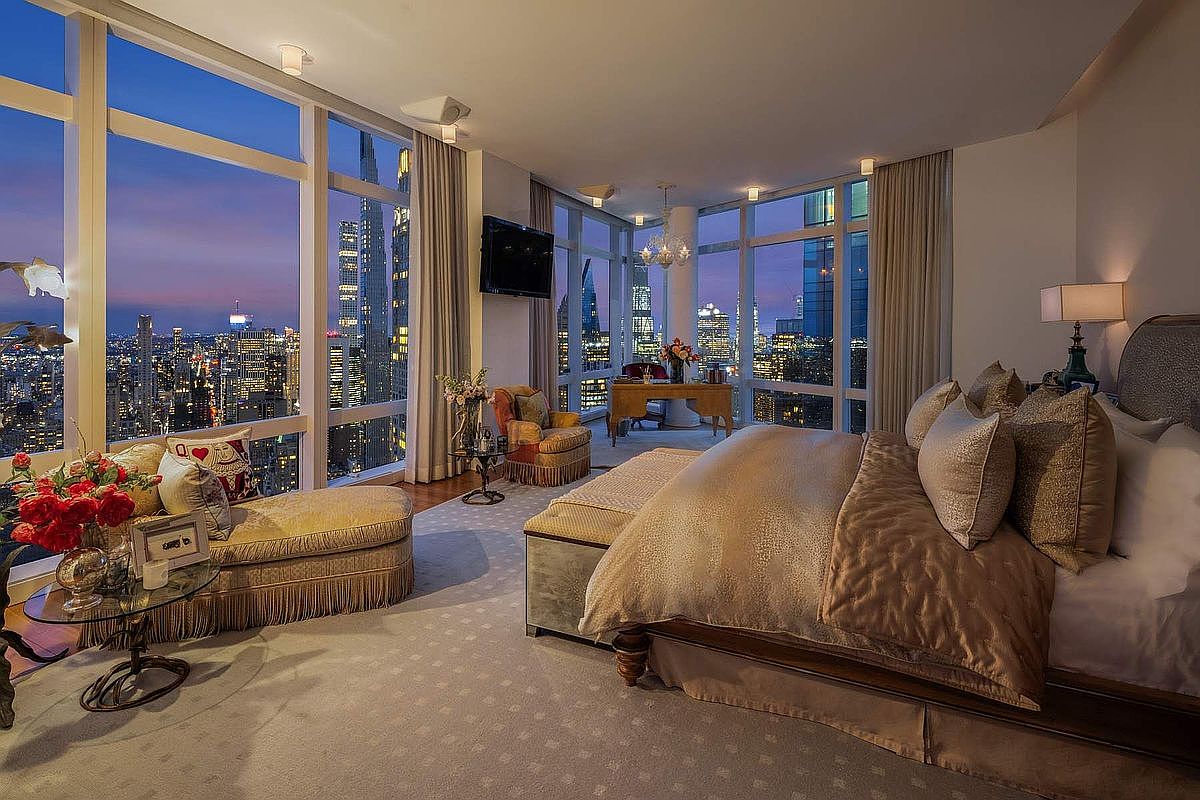 This luxurious primary bedroom features floor-to-ceiling windows that offer a breathtaking, panoramic view of the city skyline at dusk. The room is elegantly furnished with a plush king-sized bed, a sophisticated chaise lounge, and a dedicated workspace, all tied together by a neutral color palette and high-end finishes. The cinematic perspective captures the seamless blend of opulent interior design and the vibrant urban landscape outside.