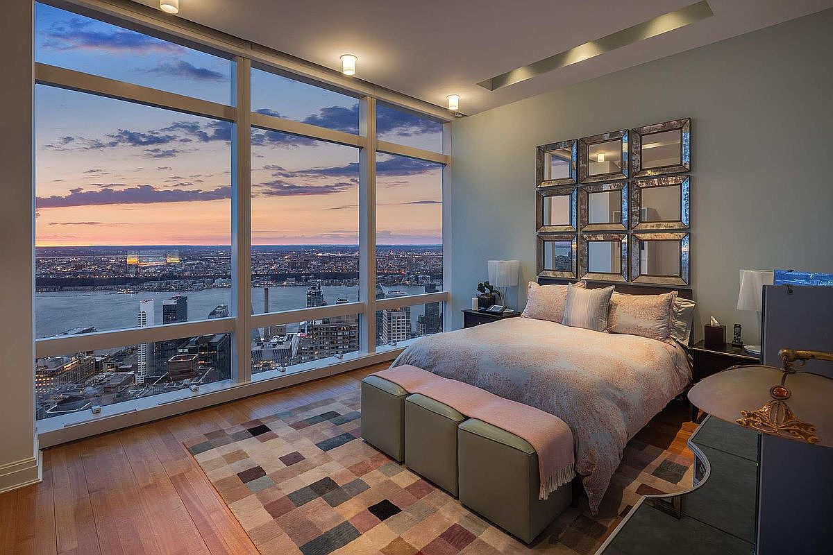 This luxurious primary bedroom features floor-to-ceiling windows that offer a breathtaking, panoramic sunset view of the city skyline and river. The room is elegantly furnished with a plush bed, a patterned area rug, and a striking grid-style mirrored wall art piece above the headboard. The overall aesthetic is sophisticated and modern, emphasizing the high-rise urban lifestyle.