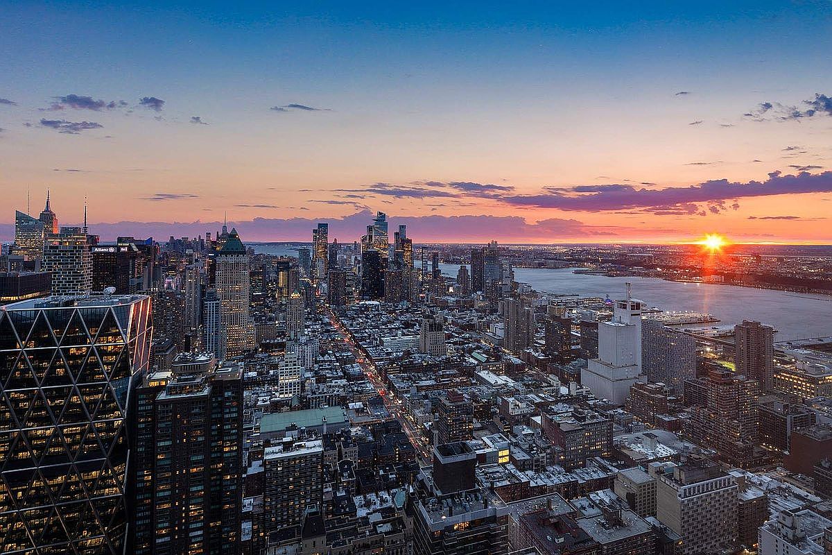 This stunning high-altitude aerial view captures the sprawling New York City skyline at sunset, showcasing a dense urban landscape of skyscrapers and city blocks. The golden hour light reflects off the Hudson River, creating a dramatic and cinematic atmosphere that emphasizes the prime location and scale of the city. The perspective provides a comprehensive look at the architectural density and vibrant energy of the metropolitan environment.