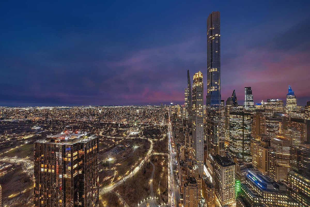 This stunning aerial view captures the iconic New York City skyline at dusk, featuring the prominent Central Park South corridor and the towering Central Park Tower. The perspective highlights the dense urban landscape, the sprawling greenery of Central Park, and the dramatic contrast between the illuminated skyscrapers and the deep purple twilight sky. It offers a high-end, cinematic look at one of the world's most prestigious real estate locations.
