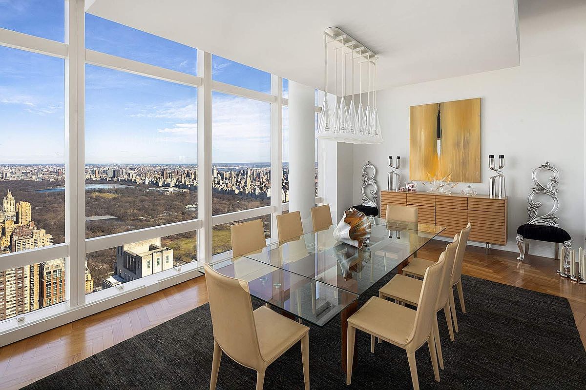 This sophisticated dining room features a sleek glass-topped table surrounded by eight modern, light-toned chairs, set atop a dark area rug. The room is defined by floor-to-ceiling windows that offer a breathtaking, panoramic aerial view of Central Park and the surrounding city skyline. A contemporary chandelier hangs above the table, while a wooden sideboard and ornate silver accent chairs add a touch of luxury to the bright, minimalist space.