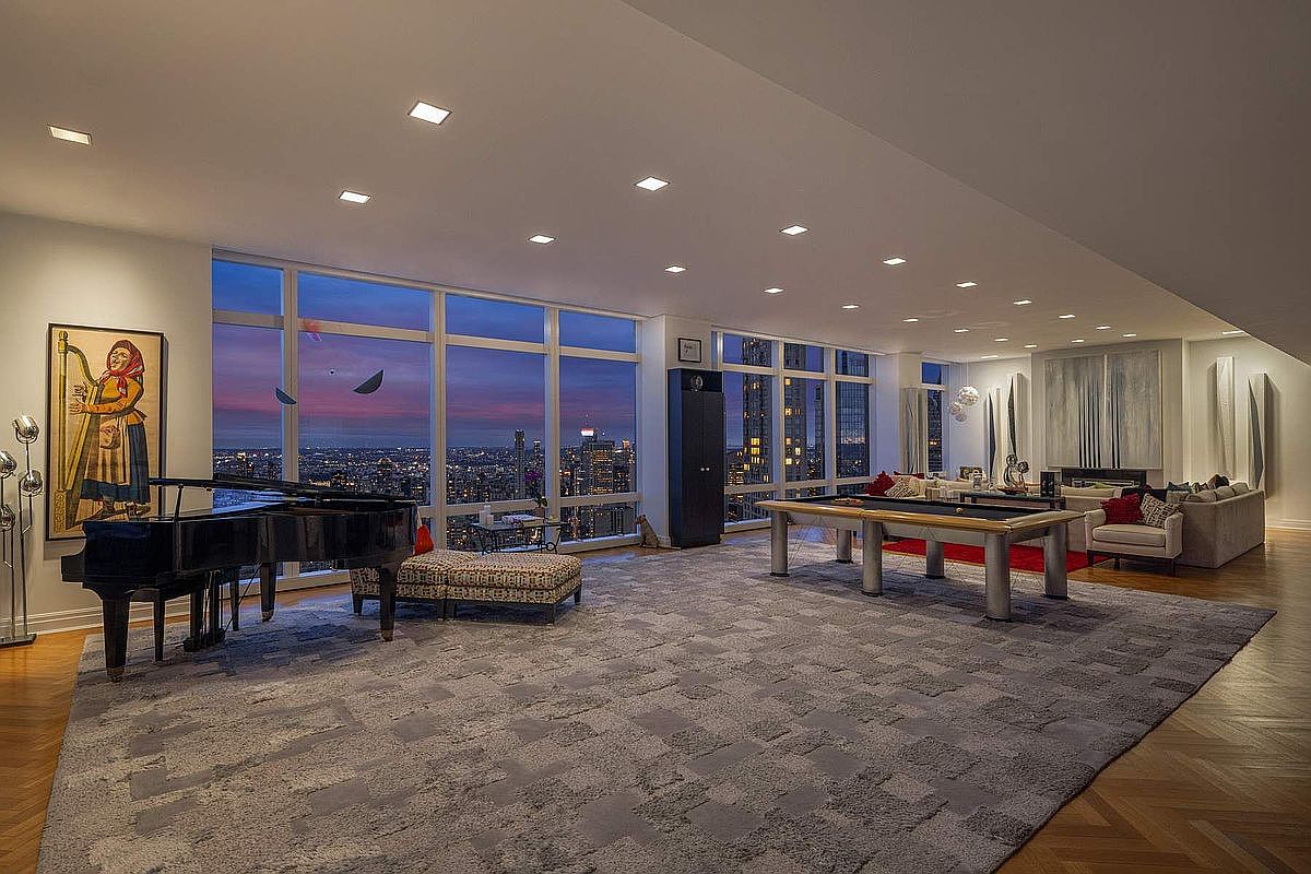 This expansive, high-rise living area features a sophisticated open-concept layout with floor-to-ceiling windows offering a breathtaking panoramic view of the city skyline at dusk. The room is elegantly furnished with a grand piano, a modern pool table, and a plush seating area, all anchored by a large, textured gray area rug over herringbone hardwood floors. The space exudes luxury and entertainment, with recessed lighting and contemporary art pieces enhancing the refined, upscale atmosphere.