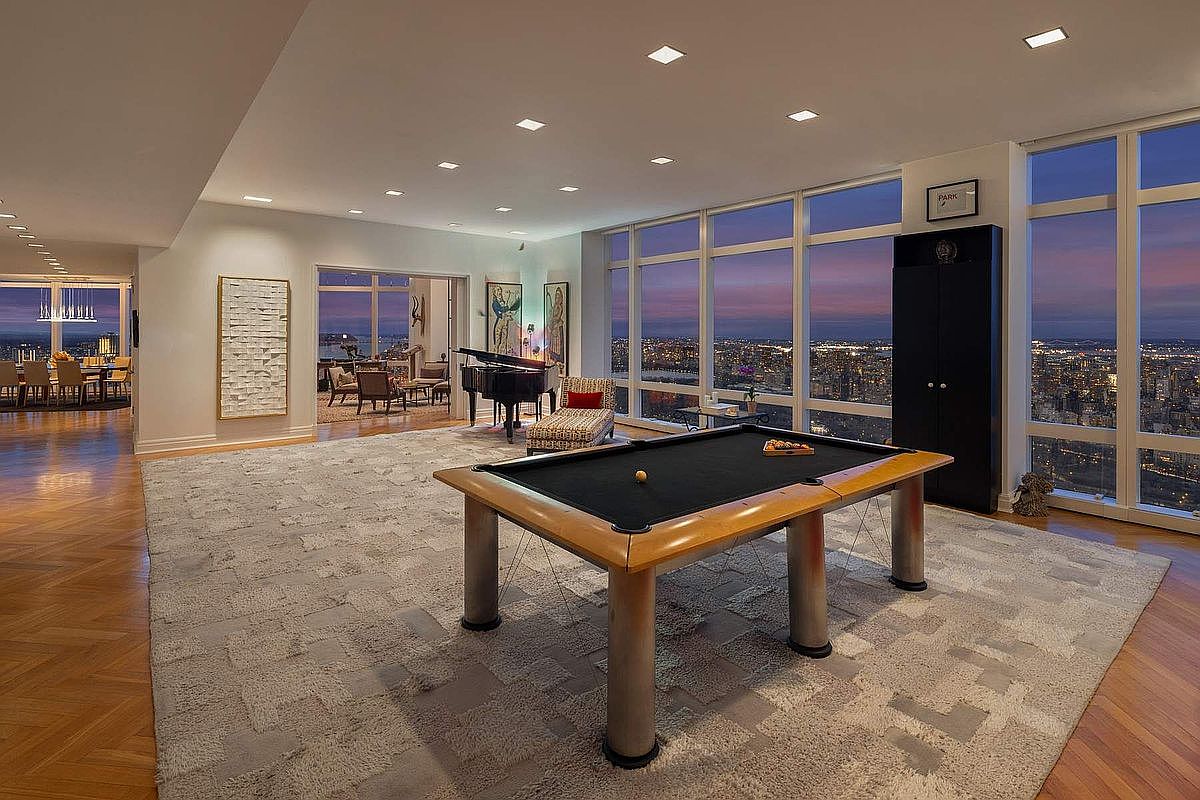 This expansive, high-rise living area features a sophisticated game room setup with a prominent pool table as the central focal point. The room is defined by floor-to-ceiling windows that offer a breathtaking, panoramic view of the city skyline at dusk, complemented by a grand piano and elegant, contemporary furnishings. The space exudes luxury and refinement, with a large, textured area rug grounding the room and recessed lighting enhancing the modern aesthetic.
