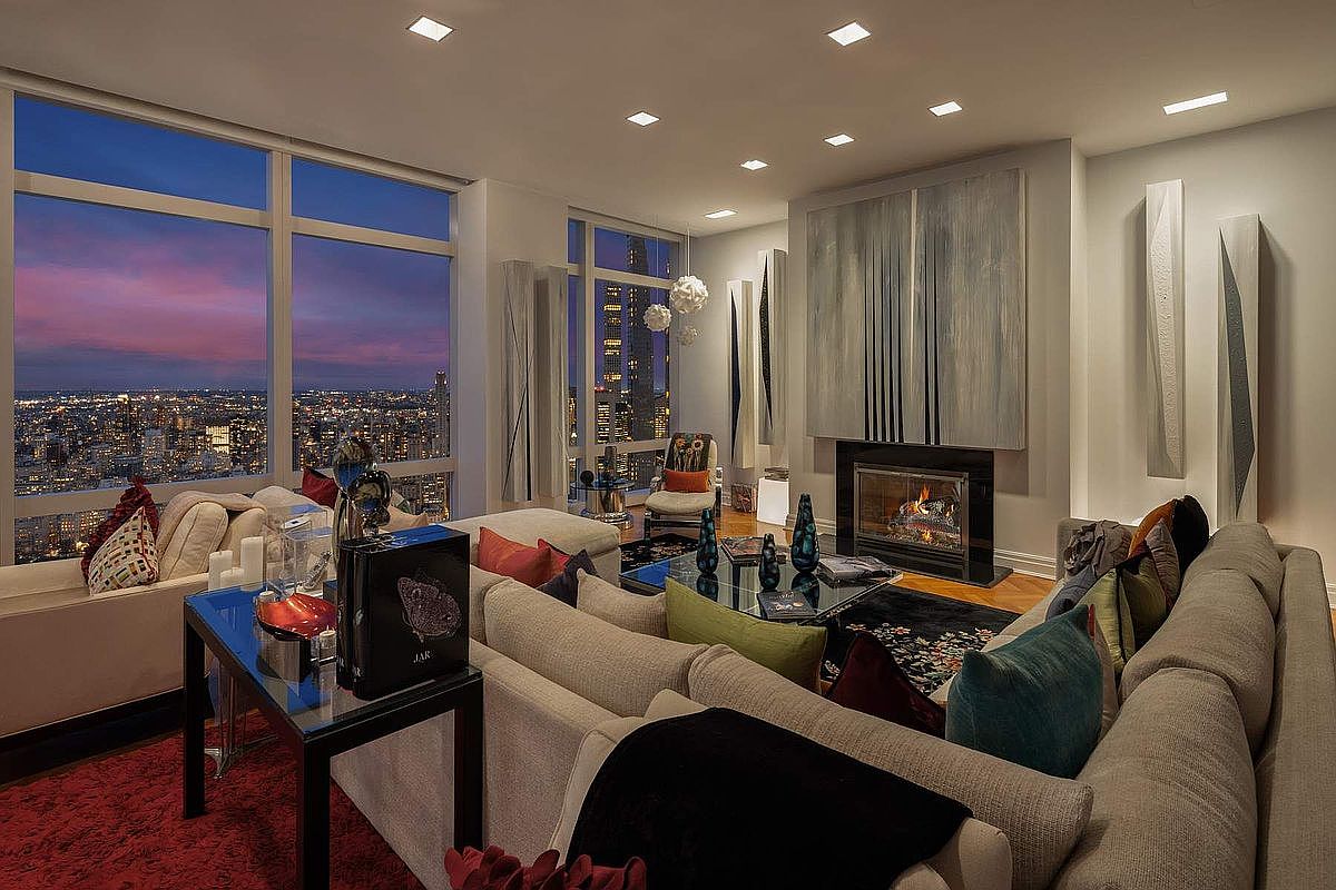 This sophisticated high-rise living room features a plush, expansive sectional sofa and a modern fireplace as its focal point, set against a backdrop of breathtaking panoramic city views at dusk. The interior design blends contemporary elegance with artistic accents, including large-scale abstract wall art and sleek glass-topped tables. The warm, ambient lighting and rich textures create an inviting, luxurious atmosphere perfect for evening relaxation.