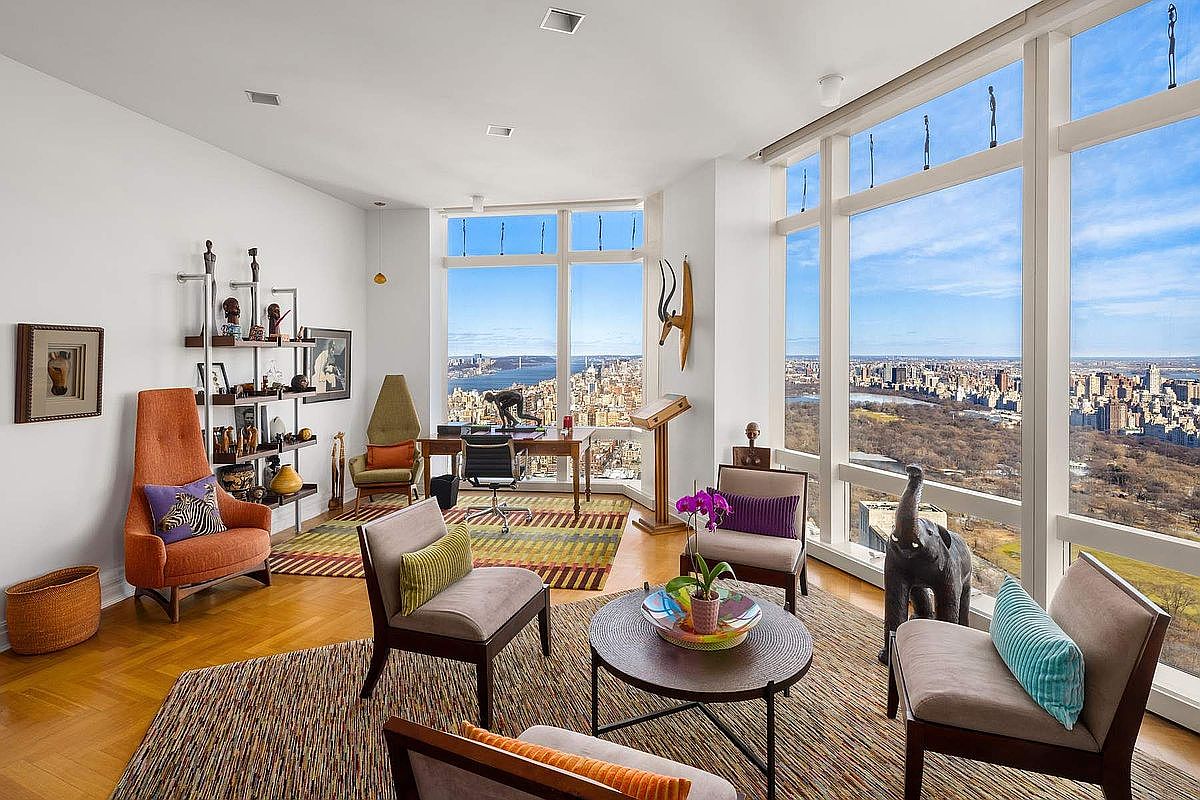 This sophisticated high-rise living room features floor-to-ceiling windows that offer a breathtaking panoramic view of the city skyline and Central Park. The space is furnished with a mix of mid-century modern seating, a circular coffee table, and a dedicated office area, all arranged on textured area rugs over hardwood floors. The room is decorated with eclectic art pieces and sculptures, creating a refined and artistic atmosphere perfect for luxury urban living.