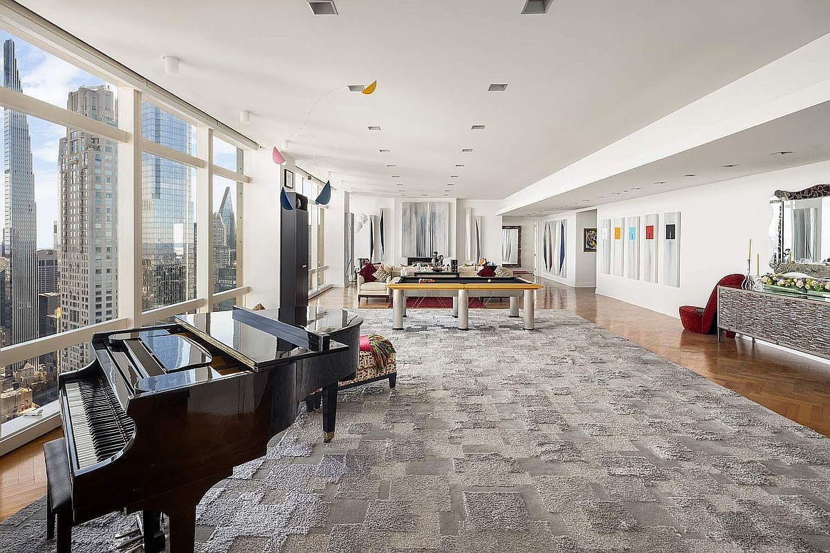 This expansive, high-floor living room features floor-to-ceiling windows offering panoramic city views, a grand piano, and a central pool table. The minimalist, modern aesthetic is defined by a large textured area rug, white walls adorned with contemporary art, and a sleek, open-concept layout. The perspective captures the vastness of the space, emphasizing its luxury, natural light, and sophisticated urban design.