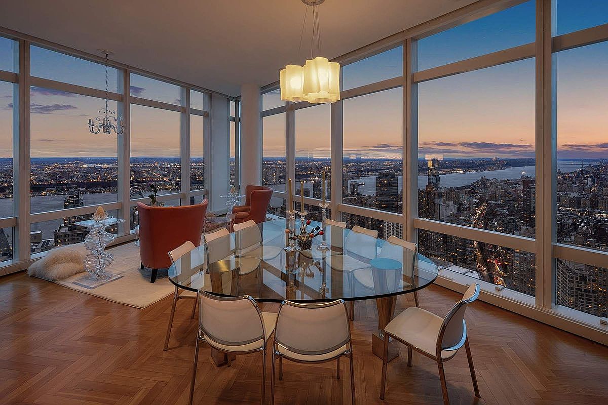 This sophisticated dining room features a sleek glass-topped table surrounded by modern white chairs, set against a backdrop of floor-to-ceiling windows offering a breathtaking panoramic view of the city skyline at dusk. The space is anchored by a unique, multi-tiered pendant light fixture and warm herringbone hardwood flooring, creating an atmosphere of refined luxury. A separate seating area with plush orange armchairs provides a cozy nook for conversation while overlooking the expansive urban landscape.