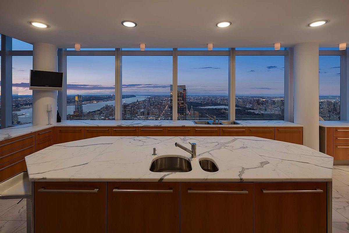 This high-end, modern kitchen features a large, central island with a striking white marble countertop and integrated sink, serving as the focal point. The space is defined by warm wood cabinetry and expansive floor-to-ceiling windows that offer a breathtaking, panoramic view of the city skyline at dusk. The clean lines and sophisticated materials create an atmosphere of luxury and urban elegance.