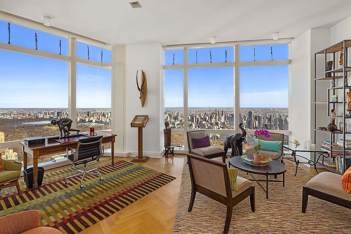 This sophisticated home office features floor-to-ceiling windows that offer a breathtaking, panoramic view of the city skyline and Central Park. The room is elegantly furnished with a wooden desk, a modern office chair, and a cozy seating area, all anchored by vibrant, patterned area rugs. The space exudes a professional yet luxurious atmosphere, perfect for high-end urban living.