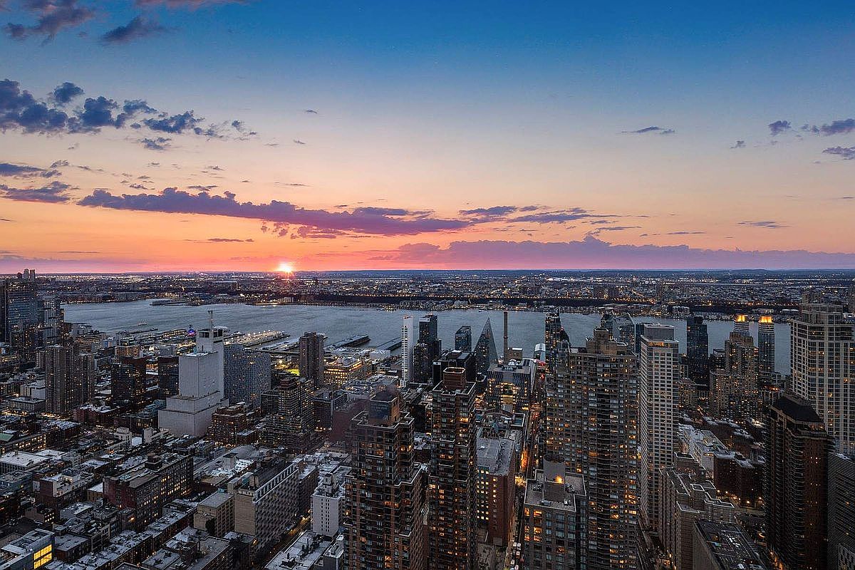This high-altitude aerial view captures a breathtaking sunset over a dense urban skyline, likely New York City, with the Hudson River reflecting the warm, fading light. The perspective emphasizes the vast scale of the city, showcasing a mix of towering skyscrapers and residential blocks illuminated by early evening lights. The composition offers a cinematic, panoramic sense of place, highlighting the vibrant metropolitan atmosphere and the dramatic transition from day to night.