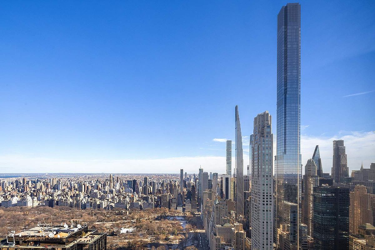 This high-altitude aerial view captures the iconic New York City skyline, dominated by the sleek, glass-clad Central Park Tower rising prominently above the surrounding skyscrapers. Below, the vast, sprawling expanse of Central Park provides a stark, natural contrast to the dense urban grid of Manhattan. The perspective offers a cinematic, sweeping look at the city's architectural density and the prestigious location of the property.