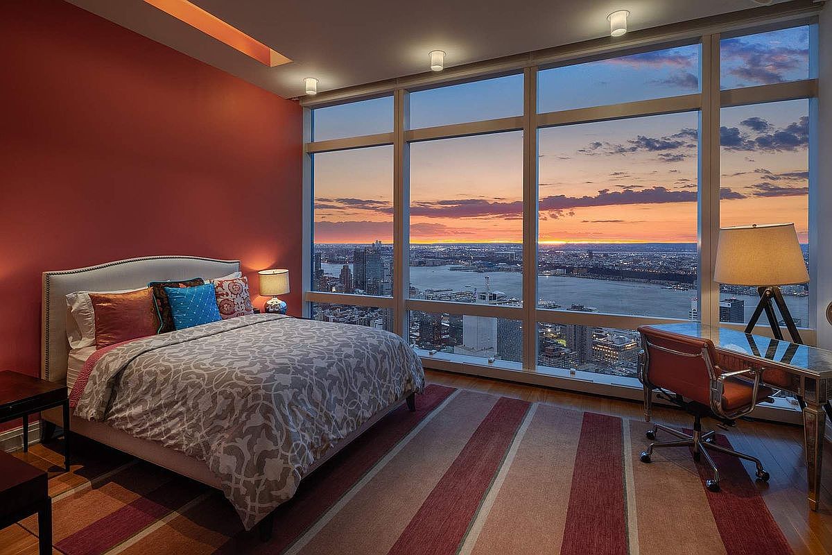 This luxurious primary bedroom features a contemporary design with a striking red accent wall and a large, patterned bed as the focal point. Floor-to-ceiling windows offer a breathtaking, panoramic sunset view of the city skyline and river, creating a dramatic and sophisticated atmosphere. A sleek glass desk and chair are positioned to take full advantage of the expansive vista, emphasizing the high-end, urban lifestyle of the property.