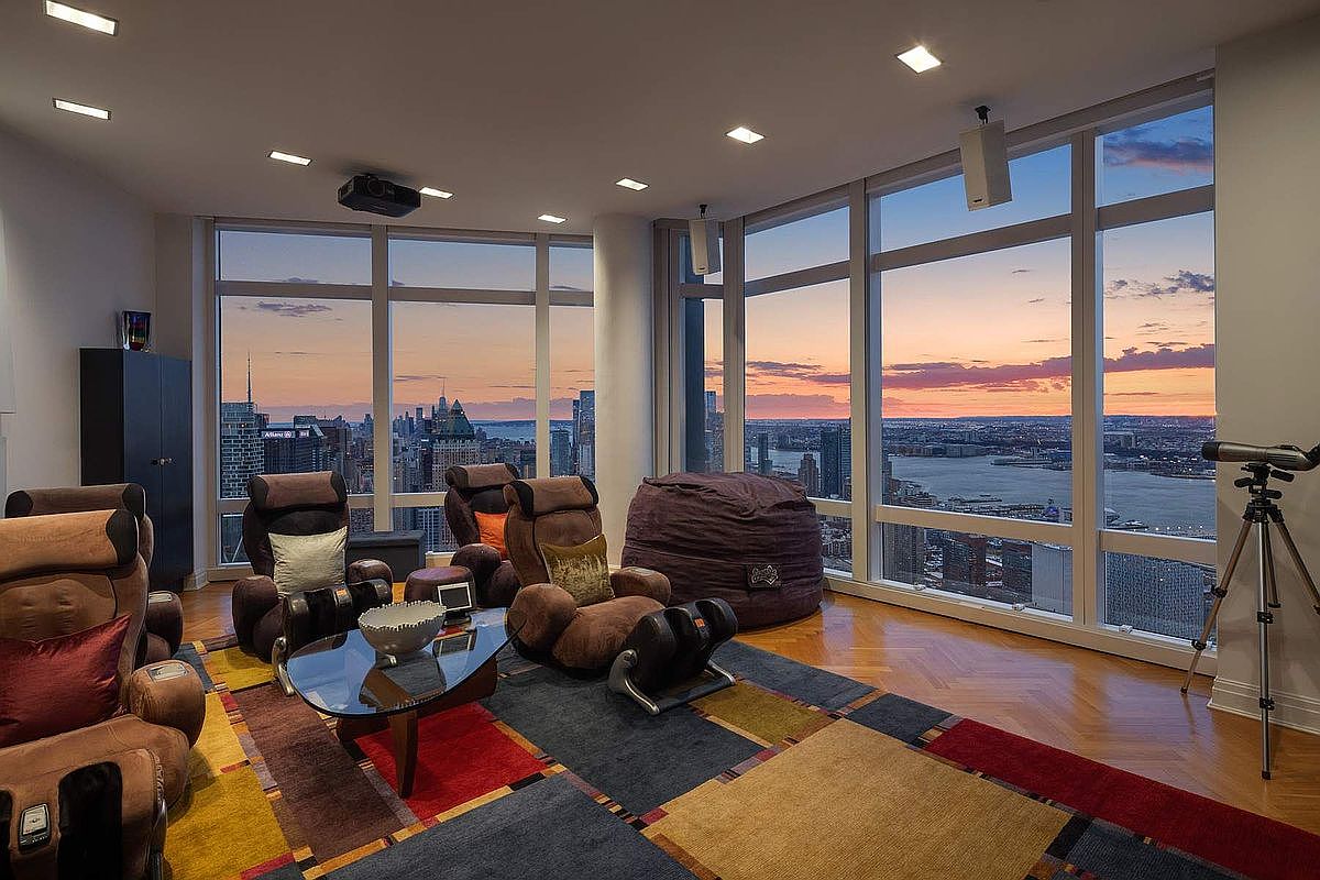 This sophisticated high-rise media room features floor-to-ceiling windows that offer a breathtaking panoramic view of the city skyline at sunset. The space is furnished with plush, ergonomic massage chairs and a large beanbag, arranged on a vibrant, multi-colored geometric area rug over hardwood floors. A projector mounted on the ceiling and a telescope positioned by the window enhance the room's function as both a cinematic retreat and an observation deck.