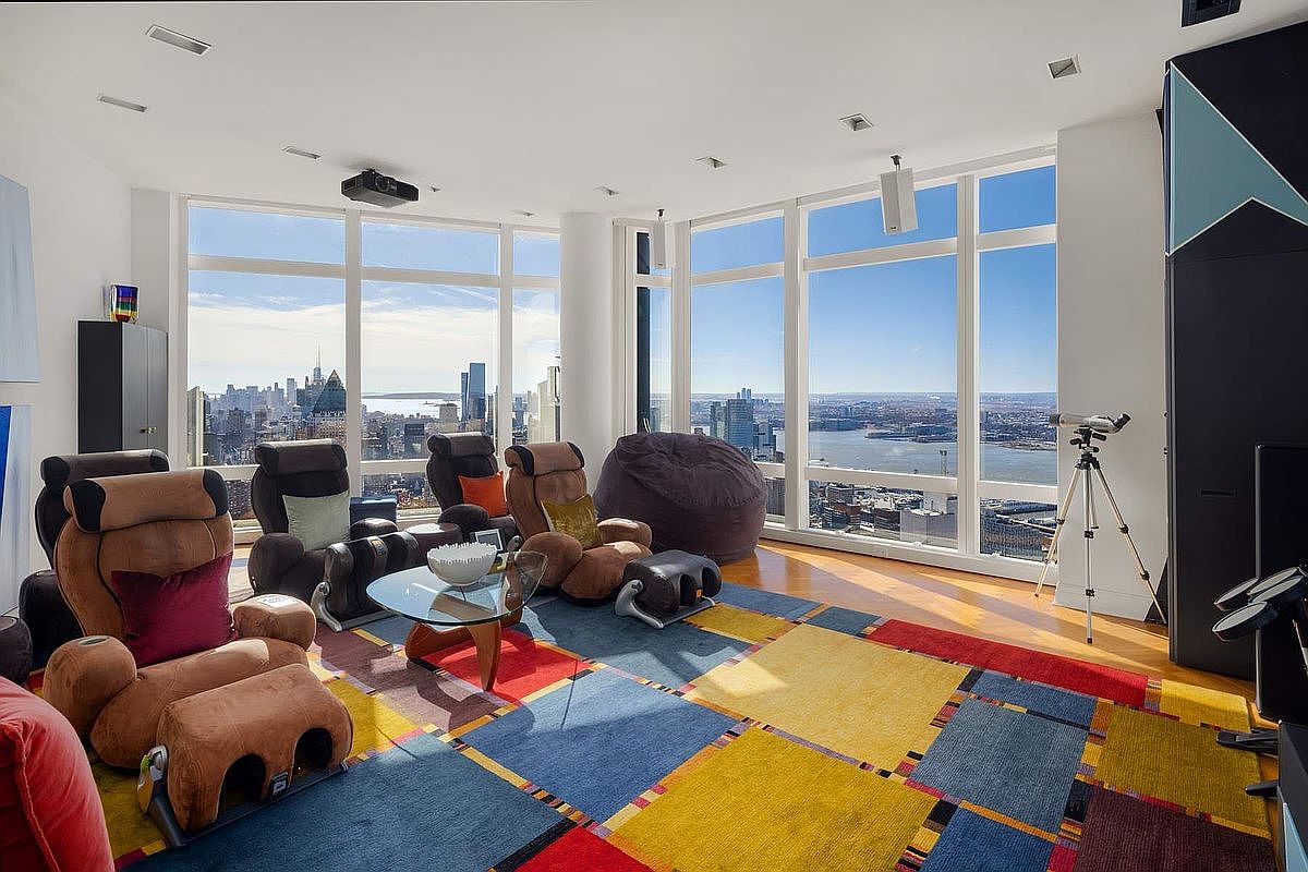 This expansive, high-rise living room features floor-to-ceiling windows that offer a breathtaking panoramic view of the city skyline and waterfront. The space is furnished with comfortable, plush massage-style recliners and a large beanbag chair, all arranged on a vibrant, multi-colored geometric area rug. A sleek glass coffee table sits in the center, while a ceiling-mounted projector suggests a dedicated media or entertainment area.