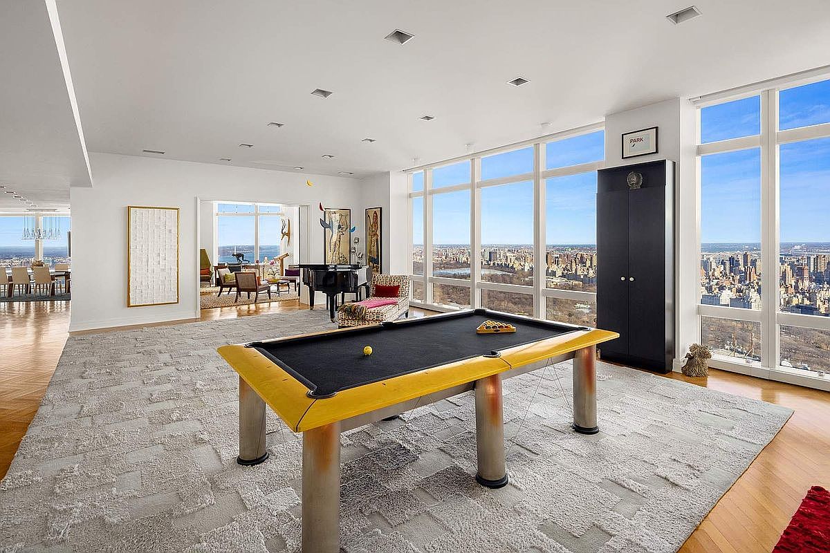 This expansive, high-floor living area features a striking yellow-trimmed pool table as its centerpiece, set against a backdrop of floor-to-ceiling windows offering panoramic city views. The room is decorated with a textured grey area rug, a grand piano, and contemporary art, creating a sophisticated and luxurious atmosphere. The open-concept layout seamlessly connects to an adjacent dining space, emphasizing the vast scale and premium nature of this urban residence.