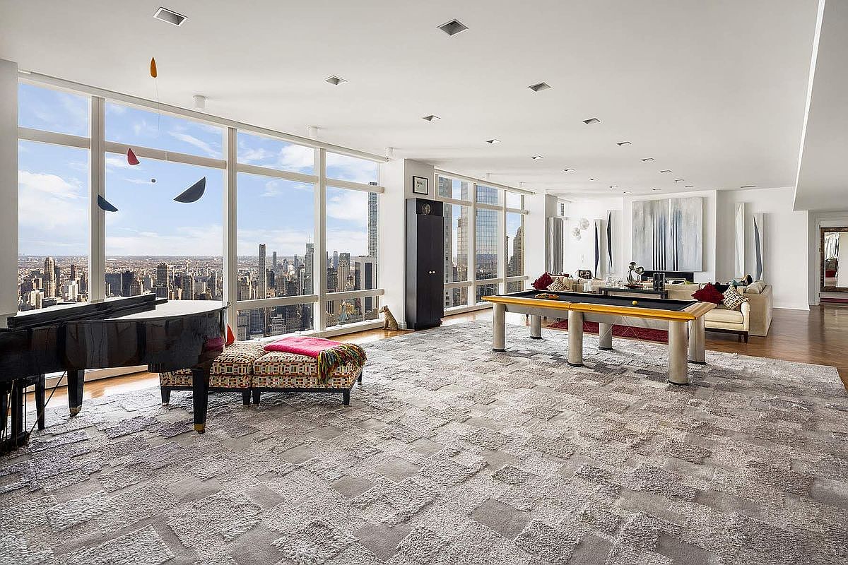 This expansive, high-floor luxury living space features floor-to-ceiling windows that offer a breathtaking panoramic view of the city skyline. The room is elegantly furnished with a grand piano, a modern pool table, and a plush, textured area rug that anchors the seating area. The minimalist, open-concept design emphasizes the dramatic scale of the room and the impressive urban backdrop.