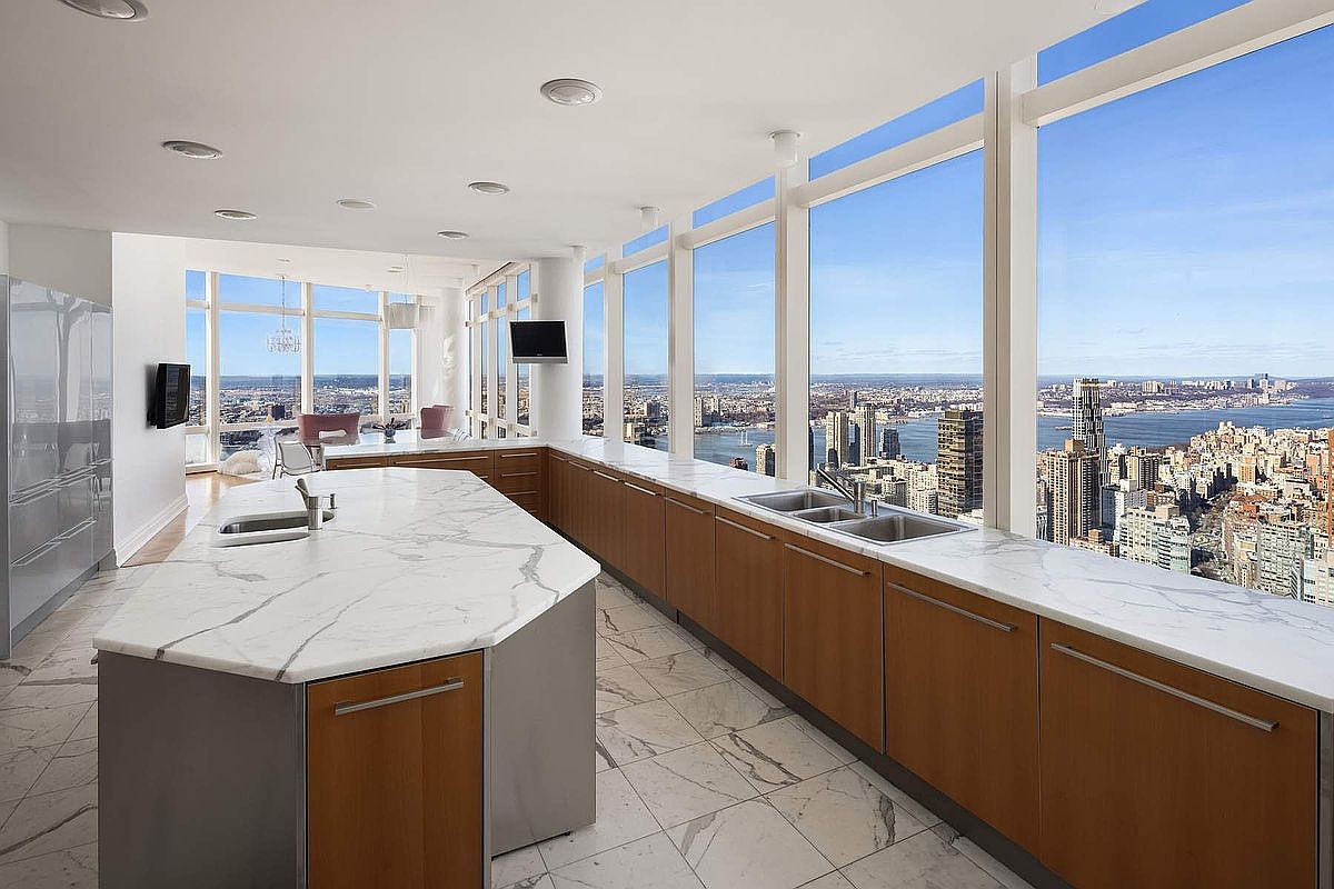 This ultra-modern, high-rise kitchen features a sleek, expansive island with a white marble countertop and warm wood cabinetry that runs along the floor-to-ceiling windows. The panoramic view of the city skyline and river provides a dramatic, cinematic backdrop, emphasizing the luxury and height of the residence. The minimalist design, combined with the bright, natural light and polished marble flooring, creates an airy and sophisticated atmosphere.
