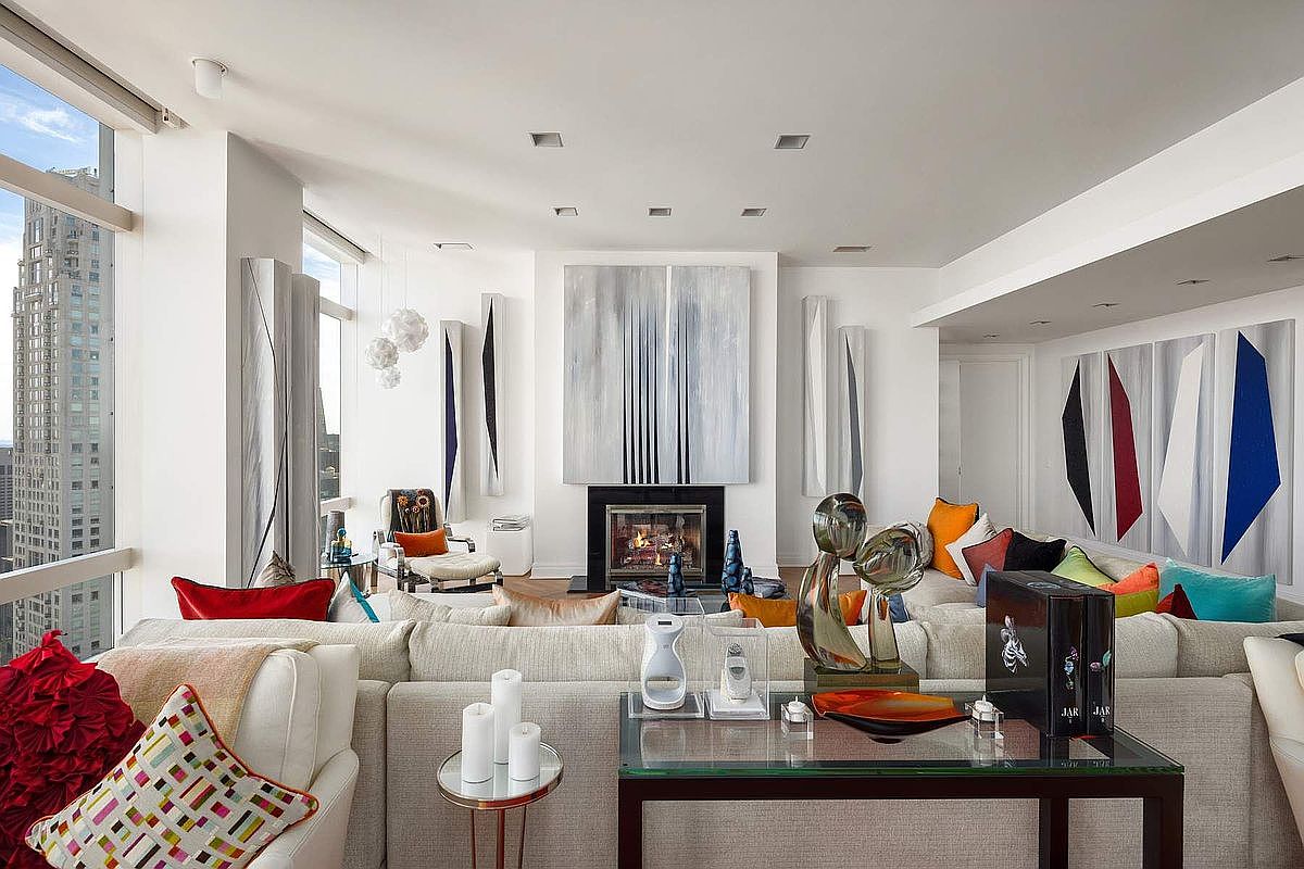 This sophisticated, high-rise living room features a bright, open-concept design with floor-to-ceiling windows offering expansive city views. The space is anchored by a central fireplace adorned with modern abstract art, complemented by a plush, neutral-toned sectional sofa accented with vibrant, colorful throw pillows. The room exudes a luxurious, contemporary aesthetic, enhanced by curated art pieces and a sleek glass-topped console table in the foreground.