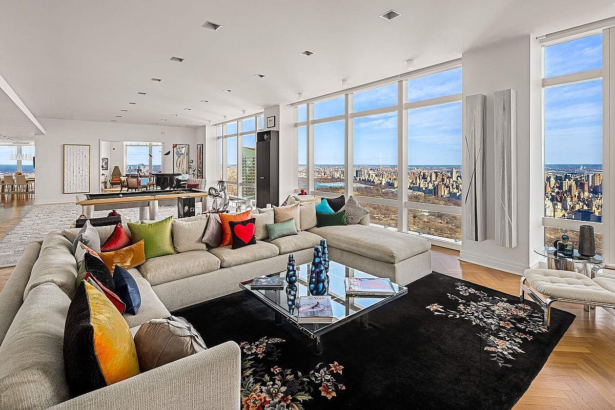 This expansive, high-floor living room features a massive L-shaped sectional sofa adorned with vibrant, multi-colored throw pillows, centered around a sleek glass coffee table on a dark floral-patterned rug. Floor-to-ceiling windows provide a breathtaking, panoramic view of the city skyline and Central Park, bathing the room in natural light. The open-concept layout seamlessly transitions into a secondary living area with a pool table, showcasing a sophisticated, modern luxury aesthetic.
