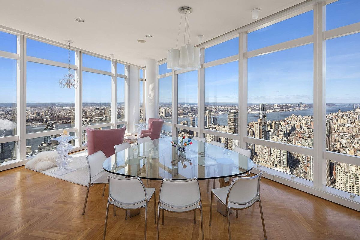 This sophisticated dining room features a large, oval glass-topped table surrounded by modern white chairs, set against floor-to-ceiling windows that offer a breathtaking, panoramic view of the city skyline and river. The space is accented by a contemporary chandelier, warm hardwood flooring in a herringbone pattern, and a cozy seating area with pink armchairs. The overall impression is one of luxury, elegance, and high-rise urban living.