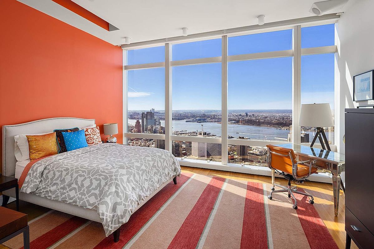 This modern primary bedroom features a striking orange accent wall and floor-to-ceiling windows that offer a breathtaking, panoramic view of the city skyline and river. The room is furnished with a comfortable bed adorned with patterned bedding and colorful throw pillows, complemented by a sleek glass-topped desk and a vibrant orange leather office chair. A striped area rug anchors the space, creating a warm and inviting atmosphere that perfectly balances urban sophistication with residential comfort.