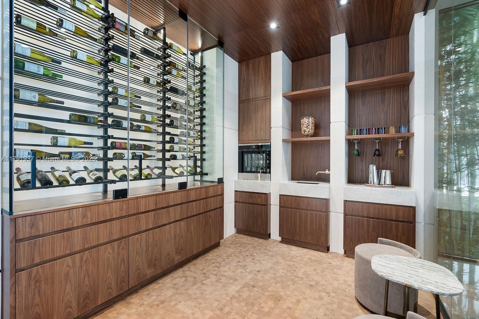 This interior shot showcases a sophisticated wine cellar, featuring a large, modern wine rack filled with bottles. The room is characterized by rich wood cabinetry and flooring, complemented by white accents and a built-in bar area with shelving and appliances. The overall design exudes luxury and functionality, perfect for wine enthusiasts.