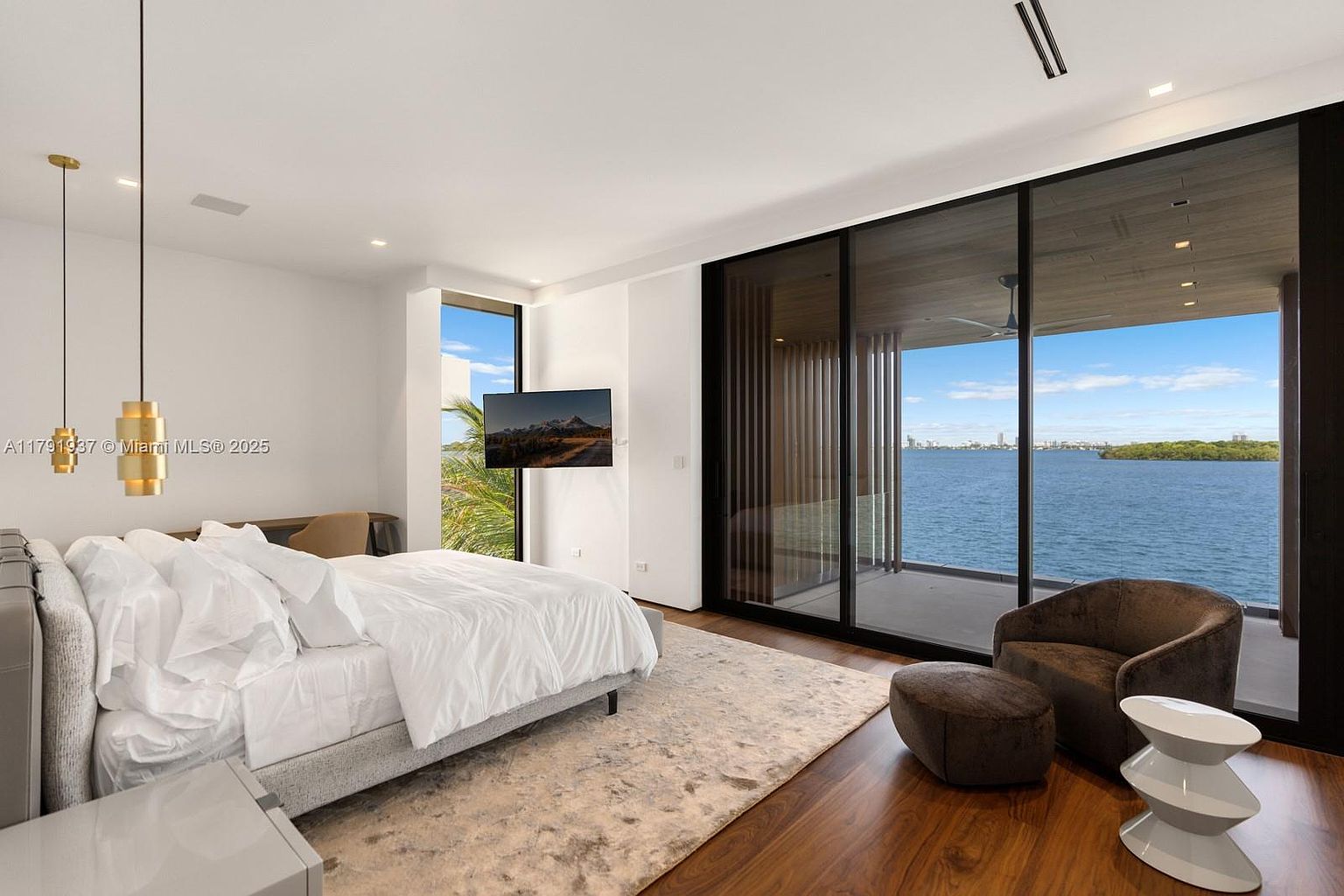 This is a luxurious primary bedroom featuring a king-size bed with white linens, a modern pendant light fixture, and a large sliding glass door that opens to a balcony with stunning water views. The room is decorated in a minimalist style with a neutral color palette, hardwood floors, and a plush area rug, creating a serene and inviting atmosphere.