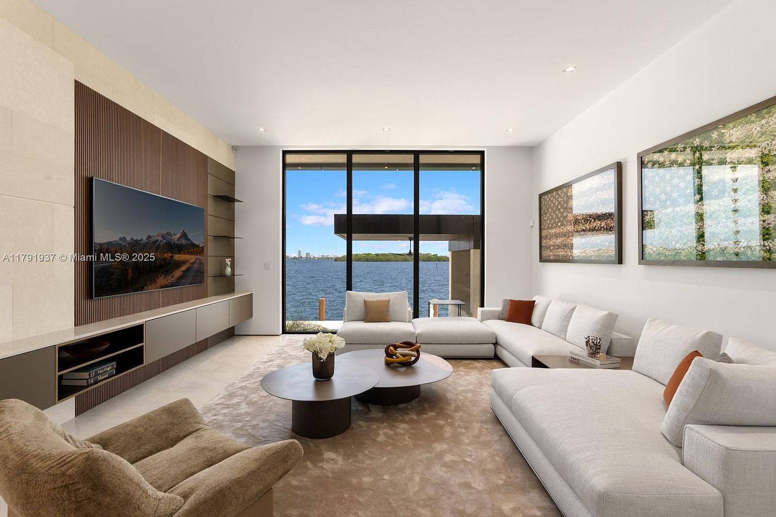 This is a bright and modern living room with a large window offering a waterfront view. The room features a sectional sofa, round coffee tables, and a media wall with a large television. The overall design is minimalist and luxurious, creating a serene and inviting atmosphere.