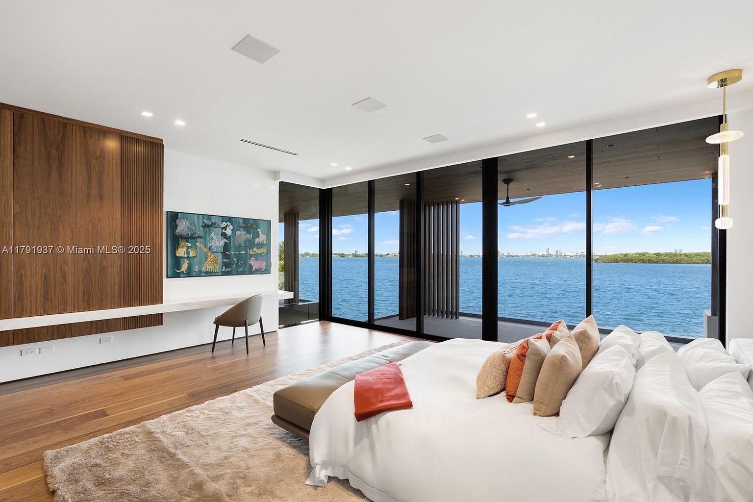This is a luxurious primary bedroom with a stunning waterfront view through large floor-to-ceiling windows. The room features a modern design with wood flooring, a large bed with plush bedding and decorative pillows, and a built-in desk area with a TV. The overall impression is one of elegance and tranquility.