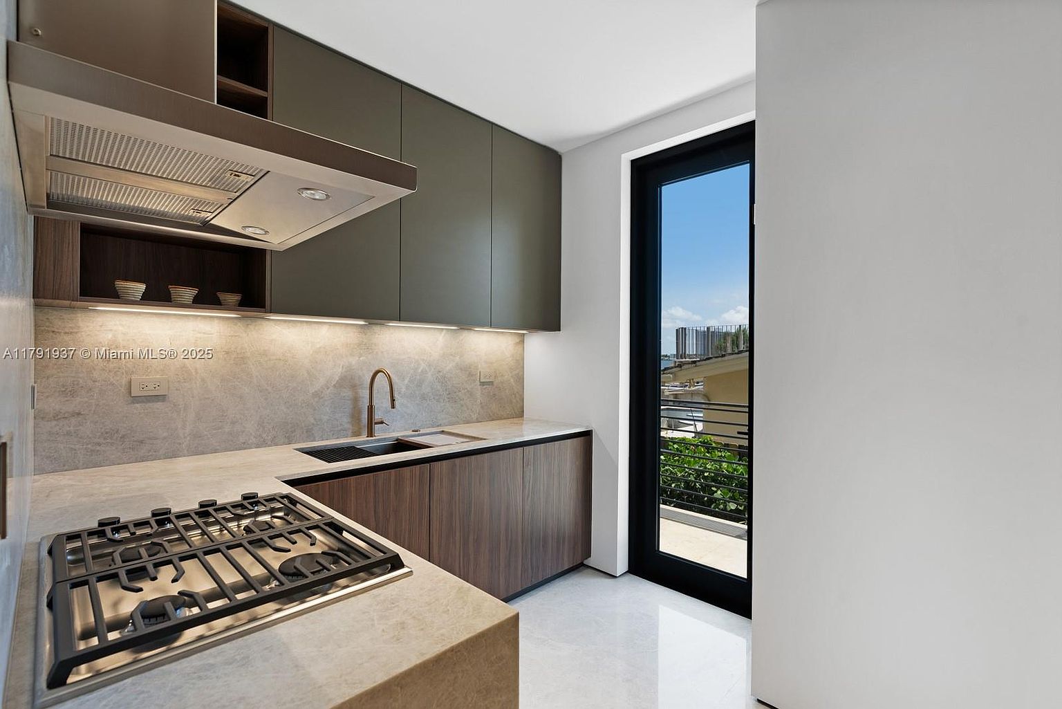This is a modern kitchen featuring sleek, dark green upper cabinets and wood-grain lower cabinets, complemented by light-colored countertops and backsplash. A stainless steel range hood is positioned above a gas cooktop, and a black-framed glass door provides natural light and a view to the outside. The overall design is minimalist and elegant.