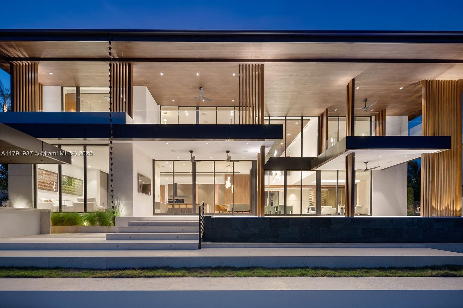 This is a striking front view of a modern, multi-level home at dusk. The architecture features a combination of wood and white elements, with large windows offering glimpses into the interior spaces. The property includes a tiered entrance with steps leading up to the main level, and the overall design exudes luxury and sophistication.