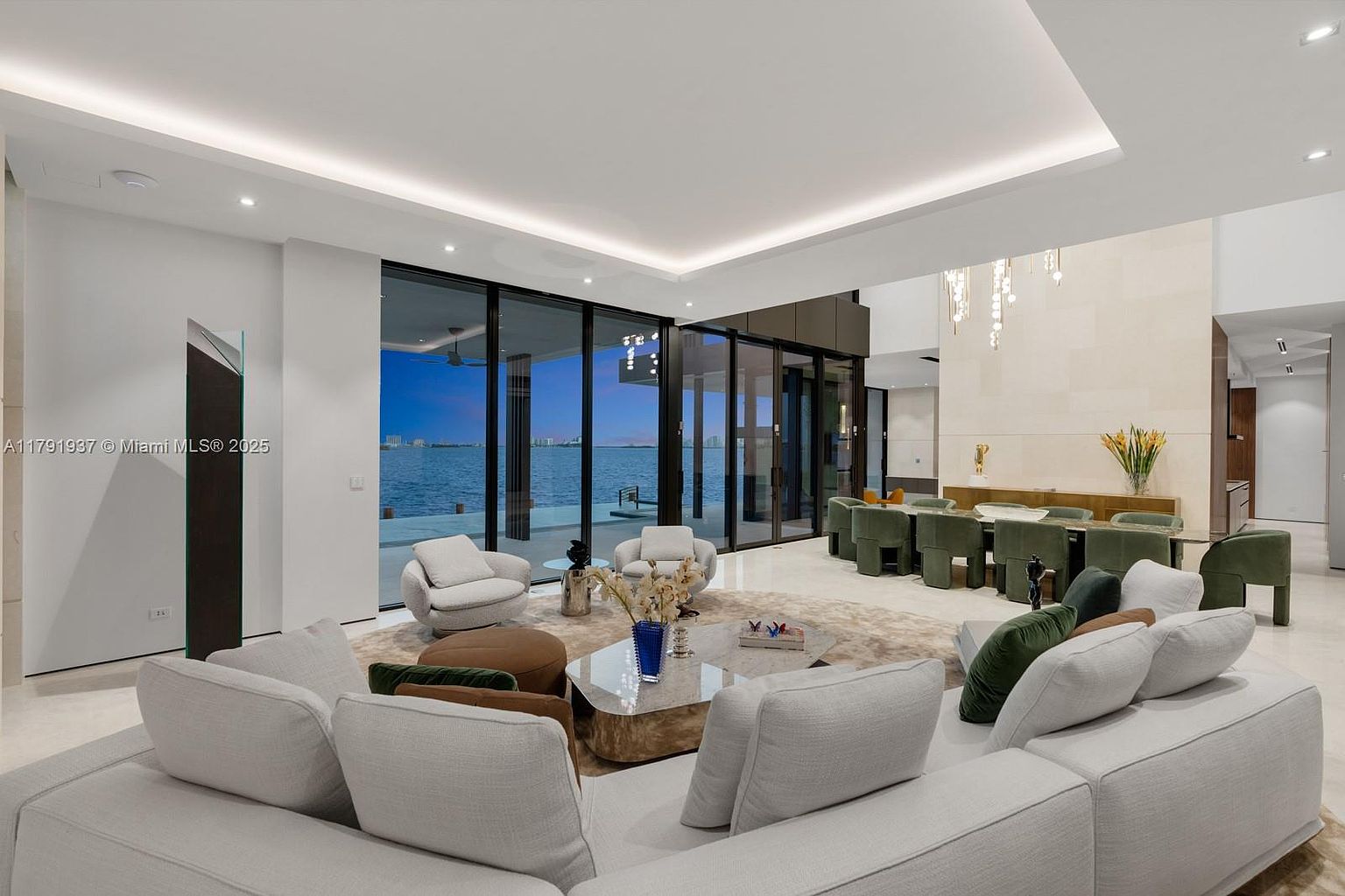 This is a spacious and luxurious living room with floor-to-ceiling windows offering a stunning waterfront view. The room features a large sectional sofa, modern armchairs, and a stylish coffee table. The open floor plan seamlessly connects the living area to the dining space, creating an inviting and sophisticated atmosphere.