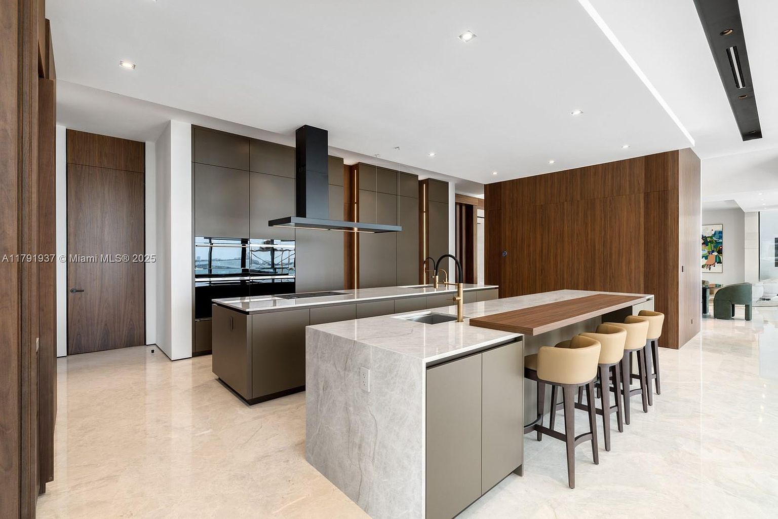 This is a wide-angle shot of a modern, high-end kitchen featuring sleek, minimalist cabinetry in a muted olive-brown tone. The kitchen boasts two islands: one with a marble countertop and sink, and the other with a wooden breakfast bar and seating for four. The space is well-lit with recessed lighting and features high-end appliances, including a modern range hood.