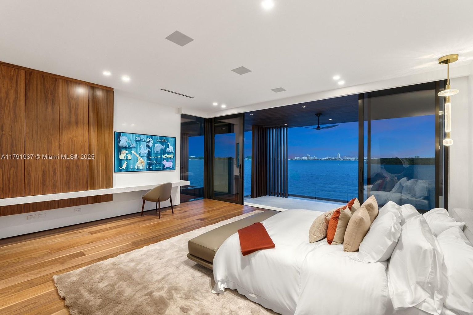 This is a luxurious primary bedroom featuring a king-size bed with white linens and decorative pillows, a wooden accent wall with a mounted TV, and a sliding glass door that opens to a waterfront view. The room has hardwood floors, a neutral-toned rug, and modern lighting fixtures, creating a serene and upscale atmosphere. The perspective is from the foot of the bed, looking towards the water view.