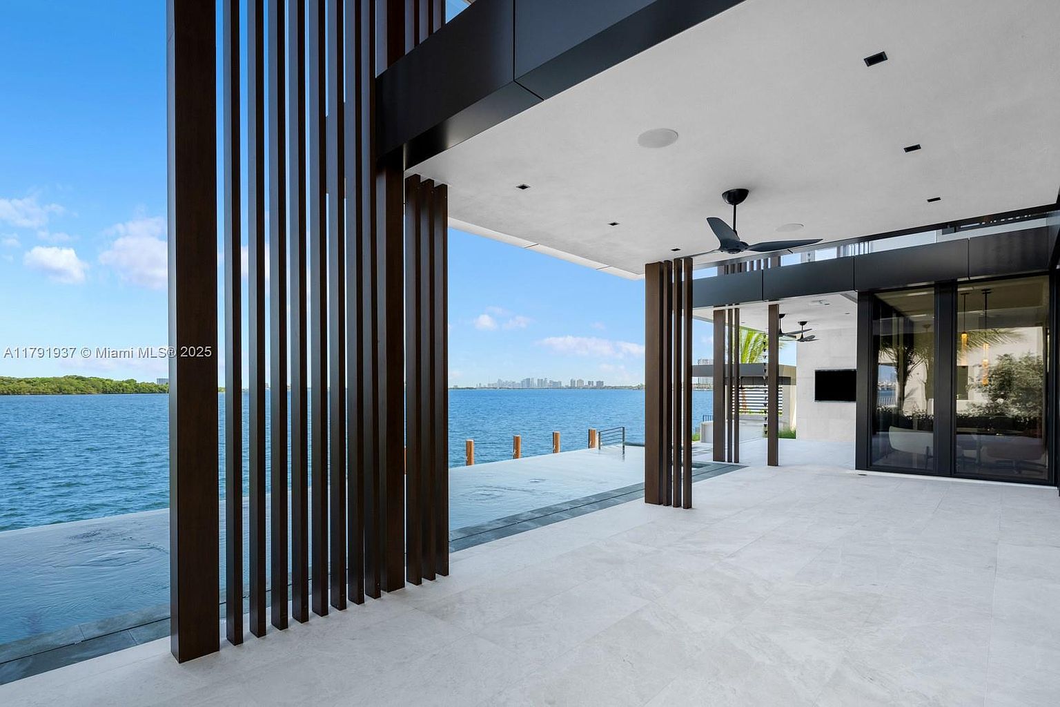 This image showcases a luxurious outdoor patio or balcony area with a stunning waterfront view. The space features sleek, modern design elements, including dark wooden vertical slats, light-colored tile flooring, and a seamless transition to an infinity pool. The open layout provides an inviting atmosphere for relaxation and entertainment, with a glimpse of the city skyline in the distance.