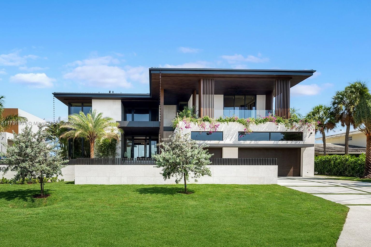 This is a front exterior view of a modern, two-story home. The house features a combination of concrete and wood elements, with a flat roof and large windows. The front yard is well-manicured with a green lawn and young trees, and a concrete wall provides privacy. The perspective is a medium shot, capturing the full facade of the house and its immediate surroundings.