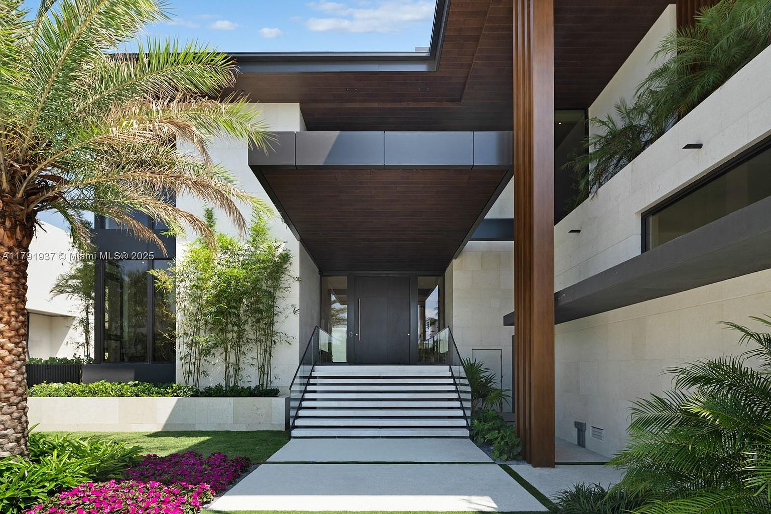 The image showcases the grand entryway of a modern residence, featuring a striking combination of natural elements and architectural design. A set of stairs with black and white accents leads to a dark-colored front door, framed by a wooden overhang and a lush landscape of palm trees and greenery. The overall impression is one of sophisticated elegance and inviting curb appeal.