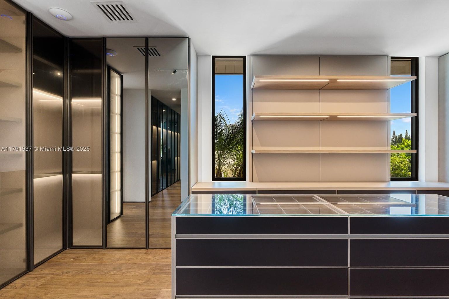 This is a modern walk-in closet featuring sleek, dark-framed glass doors and built-in shelving with integrated lighting. A central island with a glass top and dark cabinetry provides additional storage and display space. The closet has a clean, minimalist design with a focus on functionality and luxury.