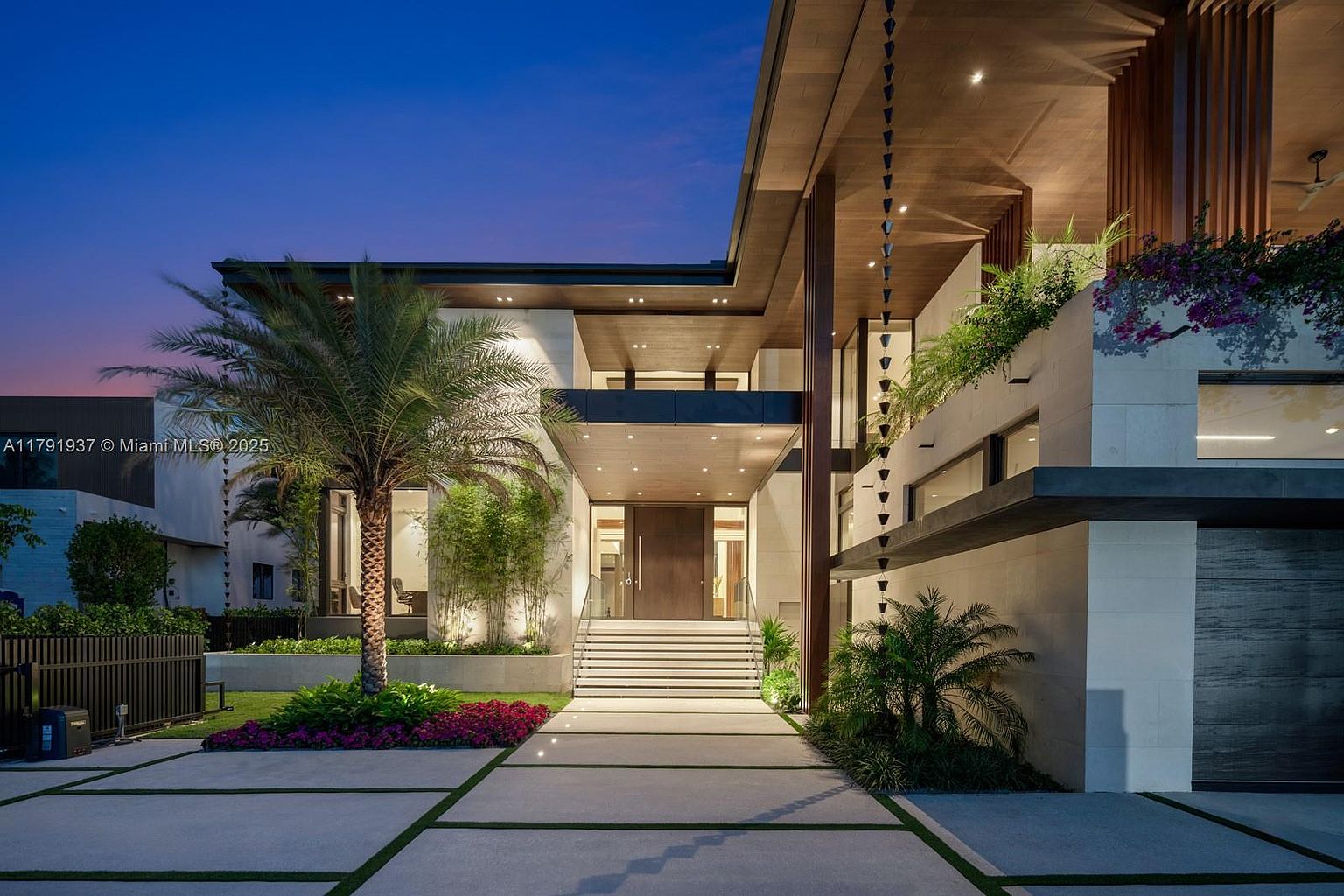 This image showcases the grand entryway of a modern luxury home at dusk. The entrance features a wide set of stairs leading to a large wooden front door, flanked by lush landscaping including palm trees and flowering plants. Architectural details include a rain chain, wood accents, and integrated lighting, creating a sophisticated and inviting atmosphere.